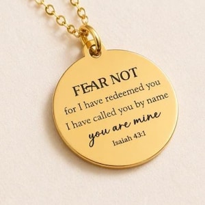 May include: A gold-toned circular pendant necklace featuring a delicate chain and a round charm engraved with the black text: FEAR NOT for I have redeemed you I have called you by name you are mine Isaiah 43:1. The piece has a polished finish.