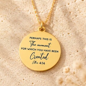 May include: A gold-colored pendant necklace with a round charm. The charm has the inscription "Perhaps this is the moment for which you have been Created" and the text "Esther 4:14". The background is a textured beige surface.