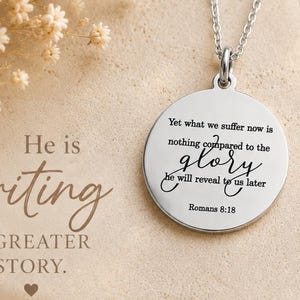 May include: A silver-toned circular pendant necklace featuring the engraved text: Yet what we suffer now is nothing compared to the glory he will reveal to us later Romans 8:18. The pendant hangs from a delicate silver chain against a neutral background.