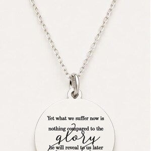 May include: A silver-toned circular pendant necklace featuring a delicate chain and a round charm engraved with the text: Yet what we suffer now is nothing compared to the glory he will reveal to us later Romans 8:18 in black lettering.