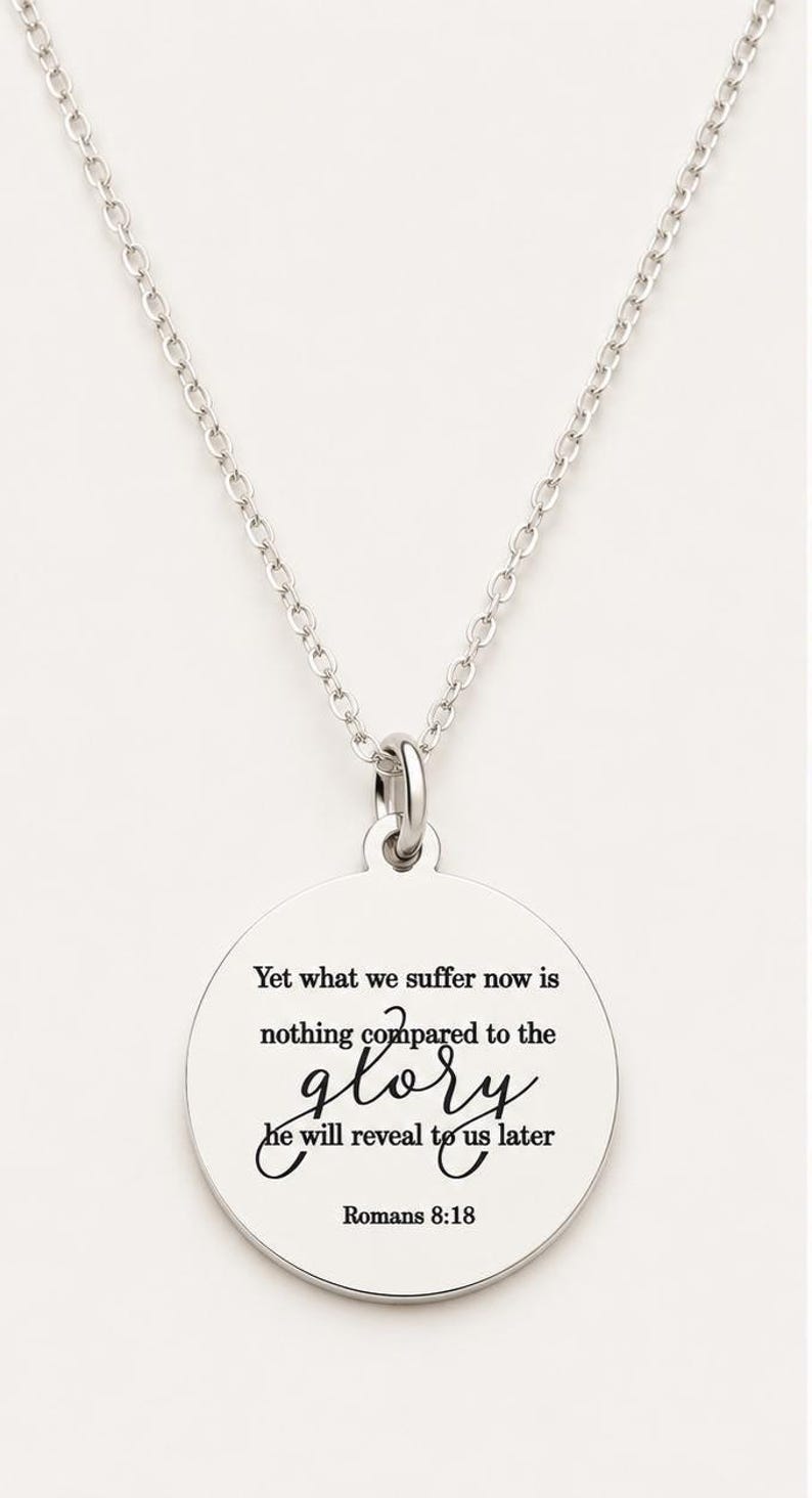May include: A silver-toned circular pendant necklace featuring a delicate chain and a round charm engraved with the text: Yet what we suffer now is nothing compared to the glory he will reveal to us later Romans 8:18 in black lettering.