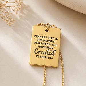 Op de afbeelding: Een goudkleurige rechthoekige hanger aan een ketting met de gegraveerde zwarte tekst: PERHAPS THIS IS THE MOMENT FOR WHICH YOU HAVE BEEN Created ESTHER 4:14. De hanger hangt aan een fijne goudkleurige ketting en heeft een gladde afwerking.