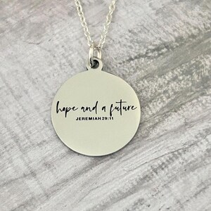 May include: A silver-colored necklace with a circular pendant. The pendant has the words "hope and a future" in a handwritten font, with "JEREMIAH 29:11" below. The chain is delicate and silver.