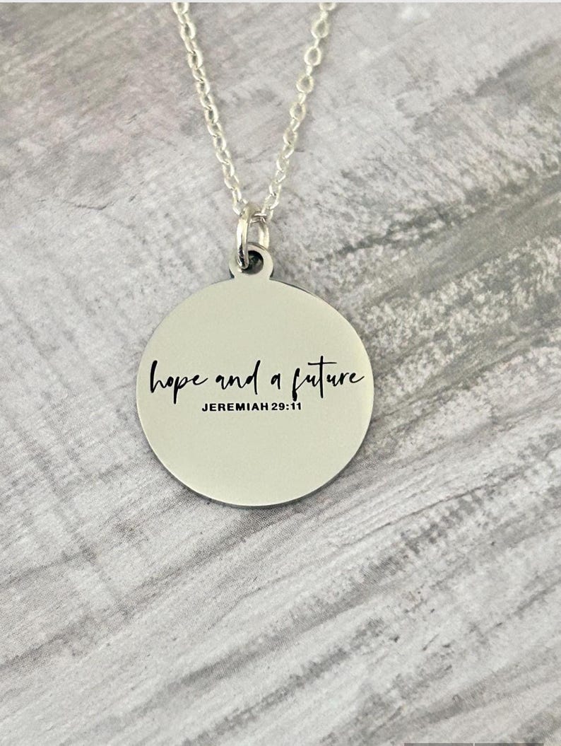 May include: A silver-colored necklace with a circular pendant. The pendant has the words "hope and a future" in a handwritten font, with "JEREMIAH 29:11" below. The chain is delicate and silver.