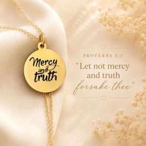 May include: A gold-colored pendant necklace with the words "Mercy and Truth" in black script. The necklace is displayed on a cream-colored fabric. The text "Proverbs 3:3" and a quote are also visible.