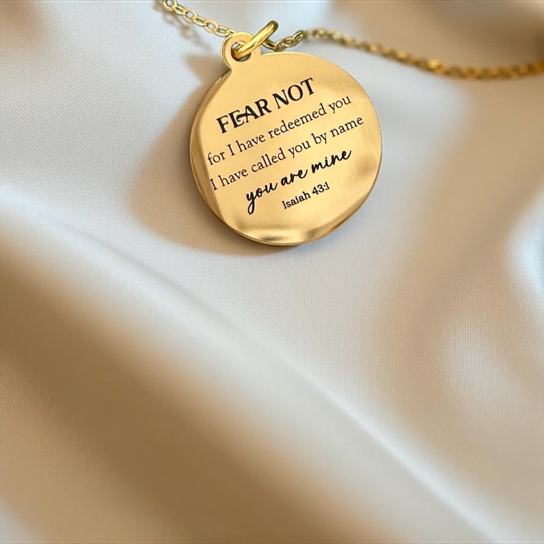 May include: A gold-toned circular pendant necklace featuring the engraved text FEAR NOT for I have redeemed you I have called you by name you are mine Isaiah 43:1. The pendant is attached to a delicate gold-toned chain with a polished finish.