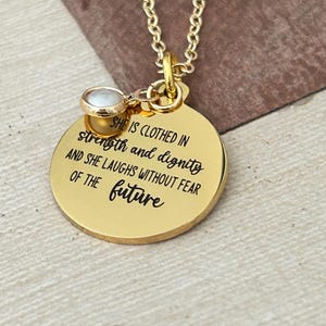 May include: Gold necklace with a round pendant that says "SHE IS CLOTHED IN strength and dignity AND SHE LAUGHS WITHOUT FEAR OF THE future". The pendant has a small pearl accent.