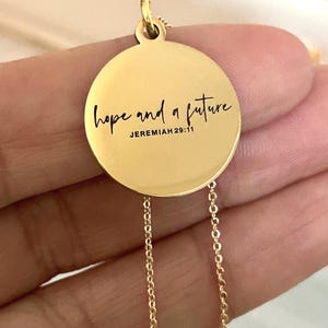 May include: Gold-colored pendant necklace with a circular charm. The charm features the words "hope and a future" in black script, with "Jeremiah 29:11" below. The necklace has a delicate chain.