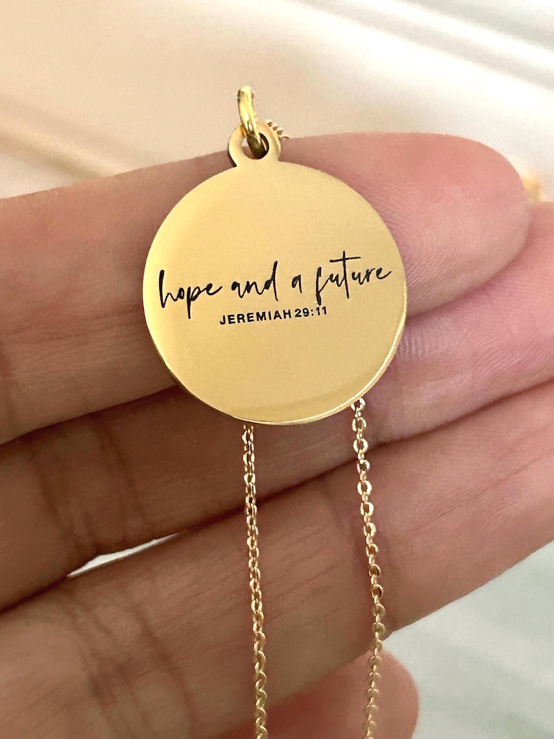 May include: Gold-colored pendant necklace with a circular charm. The charm features the words "hope and a future" in black script, with "Jeremiah 29:11" below. The necklace has a delicate chain.