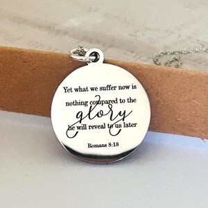 May include: A round silver-toned metal pendant necklace featuring the engraved text: Yet what we suffer now is nothing compared to the glory he will reveal to us later Romans 8:18. The pendant has a polished finish and a small circular jump ring.