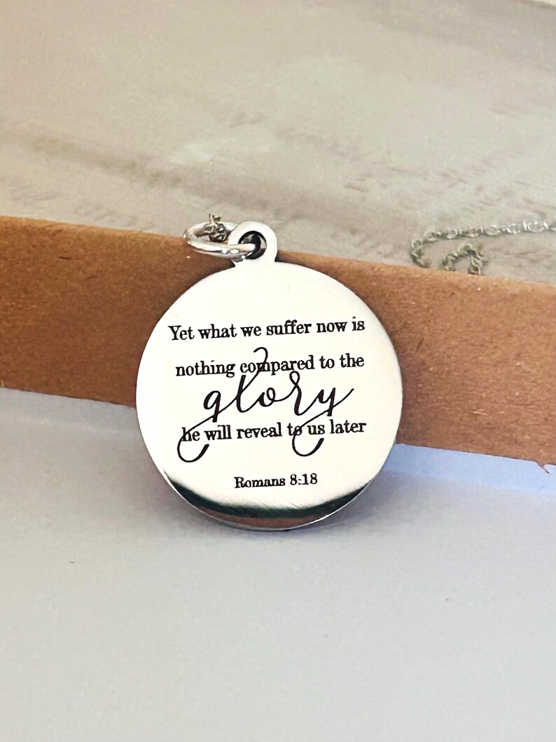 May include: A round silver-toned metal pendant necklace featuring the engraved text: Yet what we suffer now is nothing compared to the glory he will reveal to us later Romans 8:18. The pendant has a polished finish and a small circular jump ring.