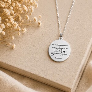 May include: A silver-toned circular pendant necklace featuring the engraved text: Yet what we suffer now is nothing compared to the glory he will reveal to us later Romans 8:18. The pendant hangs from a delicate silver-toned chain link necklace.