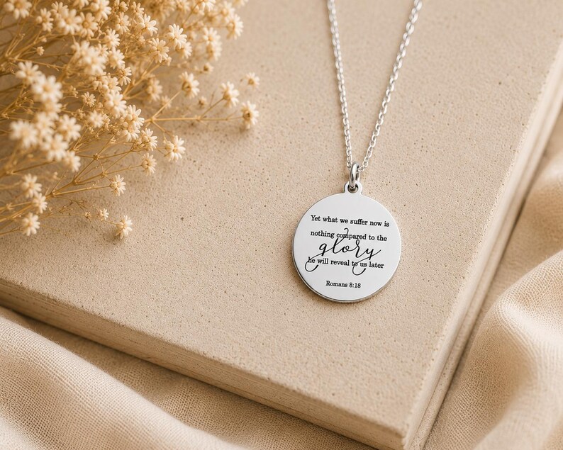 May include: A silver-toned circular pendant necklace featuring the engraved text: Yet what we suffer now is nothing compared to the glory he will reveal to us later Romans 8:18. The pendant hangs from a delicate silver-toned chain link necklace.