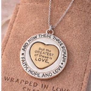 May include: Silver-tone necklace with a circular pendant featuring a gold heart and the inscription "But the GREATEST of these is LOVE." The pendant is inscribed with "Faith, Hope, and Love" and "And Now These Three Remain." The necklace is presented on a pink pouch.
