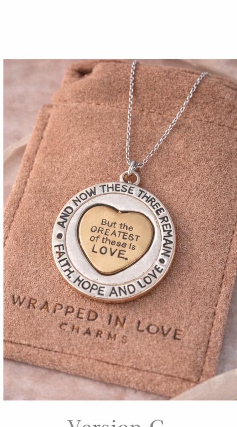 May include: Silver-tone necklace with a circular pendant featuring a gold heart and the inscription "But the GREATEST of these is LOVE." The pendant is inscribed with "Faith, Hope, and Love" and "And Now These Three Remain." The necklace is presented on a pink pouch.