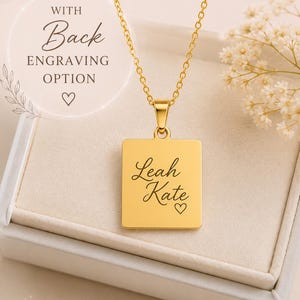 Op de afbeelding: Een goudkleurige rechthoekige hanger aan een ketting met de gegraveerde naam Leah Kate en een klein hartsymbool. De hanger hangt aan een fijne goudkleurige ketting. De tekst op de afbeelding luidt WITH Back ENGRAVING OPTION.
