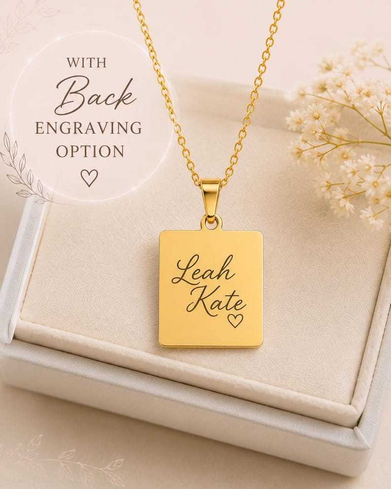 Op de afbeelding: Een goudkleurige rechthoekige hanger aan een ketting met de gegraveerde naam Leah Kate en een klein hartsymbool. De hanger hangt aan een fijne goudkleurige ketting. De tekst op de afbeelding luidt WITH Back ENGRAVING OPTION.