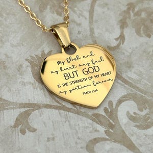 May include: A gold-colored heart-shaped pendant necklace. The pendant has an engraved inscription with the text "My flesh and my heart may fail BUT GOD IS THE STRENGTH OF MY HEART my portion forever PSALM 73:26". The necklace chain is also gold.