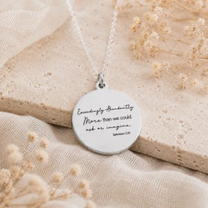 May include: A silver-coloured circular pendant necklace featuring the engraved black text Exceedingly Abundantly More than we could ask or imagine Ephesians 3:20. The pendant hangs from a delicate silver-coloured chain with a smooth, polished finish.