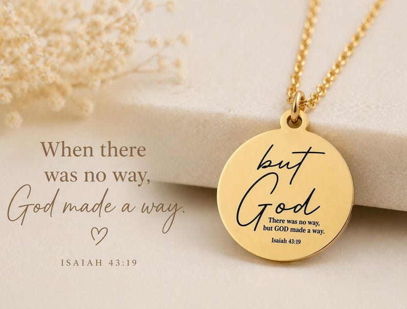 Op de afbeelding: Een goudkleurige ronde hanger aan een ketting met de gegraveerde zwarte tekst "but God" in een cursief lettertype, gevolgd door de kleinere tekst "There was no way, but GOD made a way. Isaiah 43:19" in een strak schreefloos lettertype.