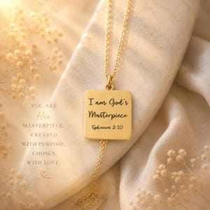 May include: A gold-colored necklace with a square pendant. The pendant is engraved with the words "I am God's Masterpiece Ephesians 2:10". The necklace is displayed on a soft, neutral-toned fabric. Additional text reads "You are His Masterpiece. Created with Purpose. Chosen with Love."
