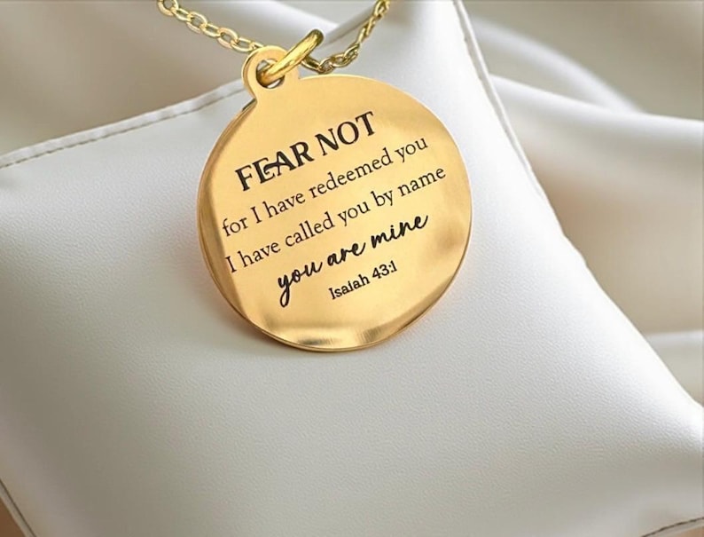 May include: A gold-toned circular pendant necklace featuring the engraved text: FEAR NOT for I have redeemed you I have called you by name you are mine Isaiah 43:1. The pendant is suspended from a delicate gold-toned chain link necklace.