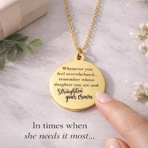 Puede incluir: Un collar con colgante dorado con un dije redondo. El dije tiene las palabras "Whenever you feel overwhelmed... remember whose daughter you are and Straighten your crown." El texto "In times when she needs it most..." también es visible.