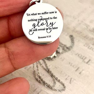 May include: A round silver-toned metal pendant featuring the engraved black text: Yet what we suffer now is nothing compared to the glory he will reveal to us later Romans 8:18. The pendant includes a small circular jump ring at the top.