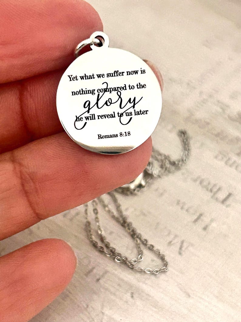 May include: A round silver-toned metal pendant featuring the engraved black text: Yet what we suffer now is nothing compared to the glory he will reveal to us later Romans 8:18. The pendant includes a small circular jump ring at the top.