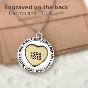 May include: A silver-toned necklace with a circular pendant. The pendant features a heart-shaped center with the text "1 COR 13:13" and is surrounded by the words "FAITH, HOPE AND LOVE" and "AND NOW THESE THREE REMAIN". The text "Engraved on the back 1 Corinthians 13:13" is also visible.