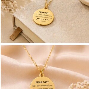 May include: A gold-toned circular pendant necklace featuring the engraved text FEAR NOT for I have redeemed you I have called you by name you are mine Isaiah 43:1. The pendant hangs from a delicate gold-toned chain with a polished metal finish.