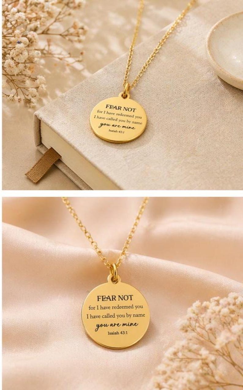 May include: A gold-toned circular pendant necklace featuring the engraved text FEAR NOT for I have redeemed you I have called you by name you are mine Isaiah 43:1. The pendant hangs from a delicate gold-toned chain with a polished metal finish.