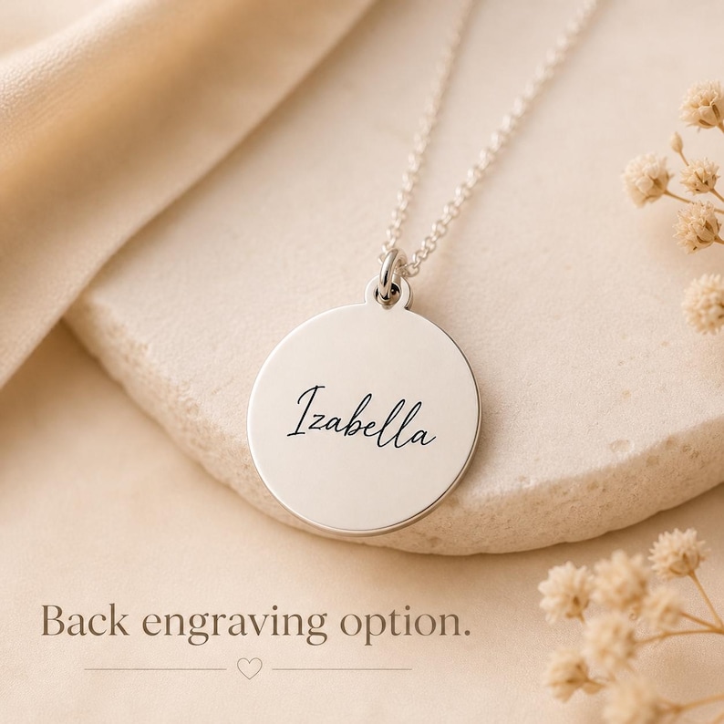 May include: A silver-colored pendant necklace with the name "Izabella" engraved on a round disc. The necklace is displayed on a neutral-toned surface with the text "Back engraving option."
