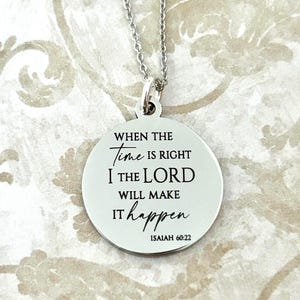 May include: A silver-colored pendant necklace with a round charm. The charm has the text "When the Time is Right I the Lord Will Make it Happen" and "Isaiah 60:22" in black lettering.
