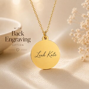 May include: A gold-toned circular pendant necklace featuring the name Leah Kate engraved in a black script font. The pendant hangs from a delicate gold-toned chain. Text to the left of the pendant reads WITH Back Engraving OPTION.