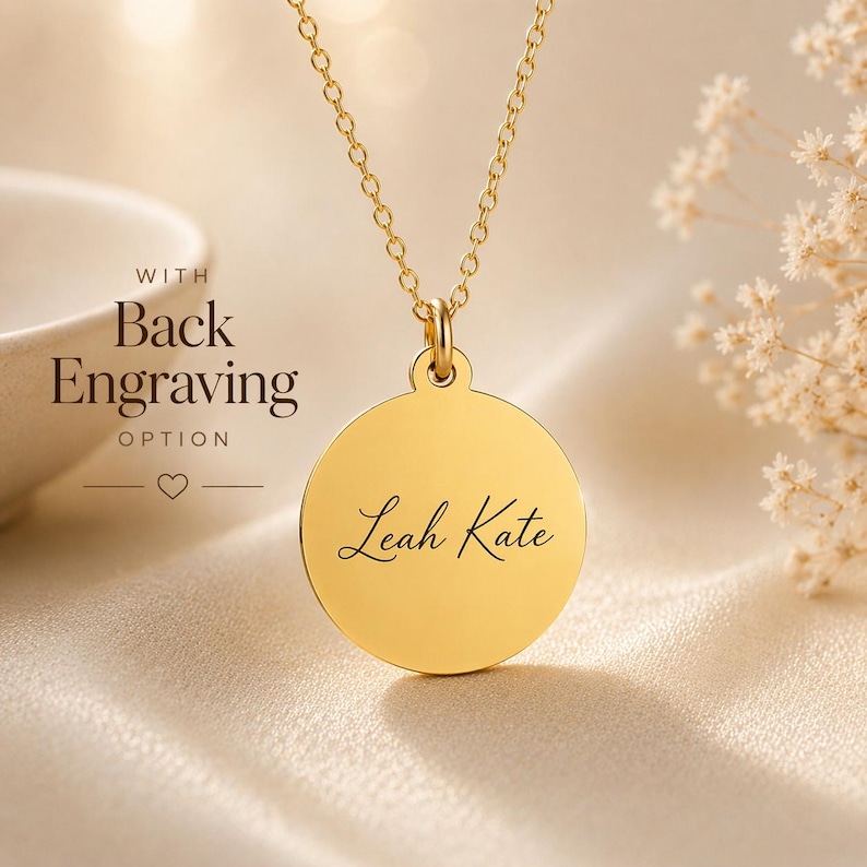 May include: A gold-toned circular pendant necklace featuring the name Leah Kate engraved in a black script font. The pendant hangs from a delicate gold-toned chain. Text to the left of the pendant reads WITH Back Engraving OPTION.