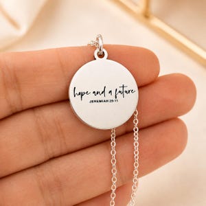 May include: A silver-colored pendant necklace with a circular charm. The charm has the words "hope and a future" in a handwritten font, with "Jeremiah 29:11" below. The necklace chain is delicate and silver.