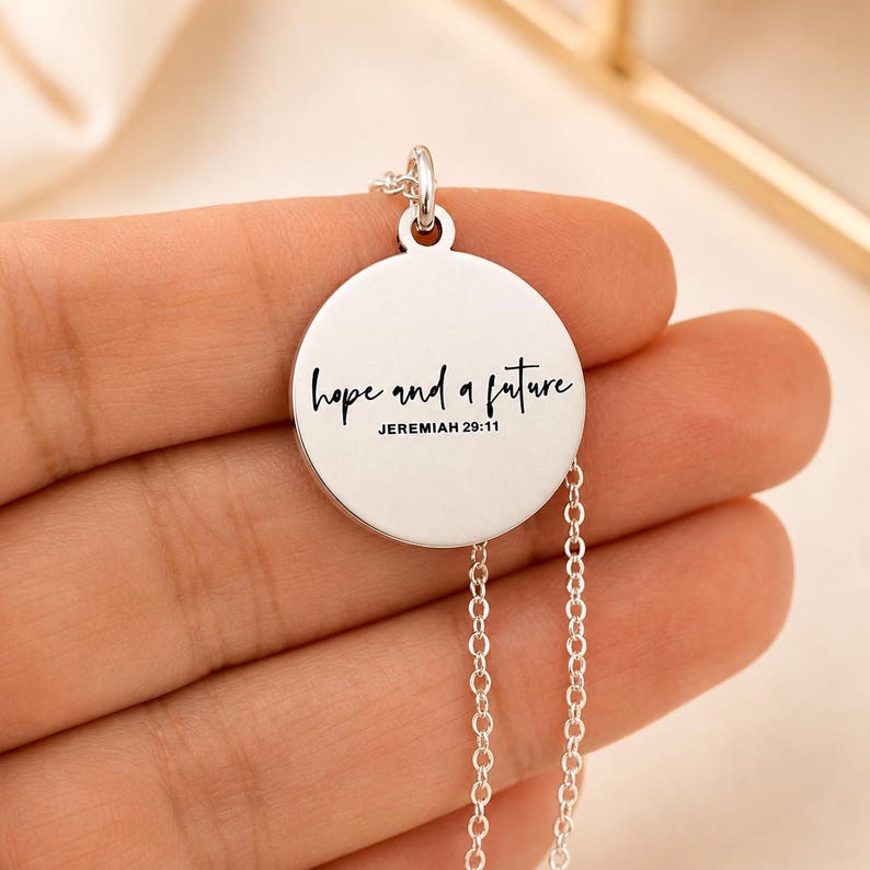 May include: A silver-colored pendant necklace with a circular charm. The charm has the words "hope and a future" in a handwritten font, with "Jeremiah 29:11" below. The necklace chain is delicate and silver.