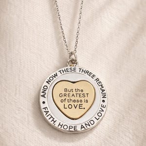 May include: Silver-toned necklace with a circular pendant. The pendant features a gold heart in the center with the words "But the GREATEST of these is LOVE." The outer ring reads "FAITH, HOPE AND LOVE" and "AND NOW THESE THREE REMAIN."