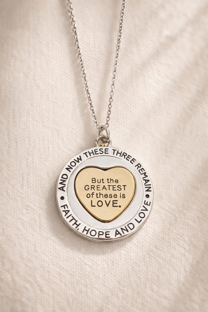 May include: Silver-toned necklace with a circular pendant. The pendant features a gold heart in the center with the words "But the GREATEST of these is LOVE." The outer ring reads "FAITH, HOPE AND LOVE" and "AND NOW THESE THREE REMAIN."