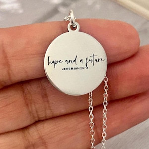 May include: A silver-colored pendant necklace with a round charm. The charm has the words "hope and a future" and "Jeremiah 29:11" in black script. The necklace has a delicate chain.