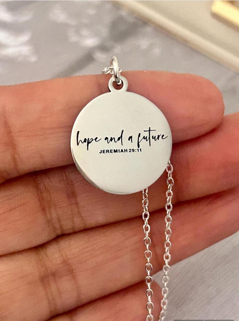 May include: A silver-colored pendant necklace with a round charm. The charm has the words "hope and a future" and "Jeremiah 29:11" in black script. The necklace has a delicate chain.