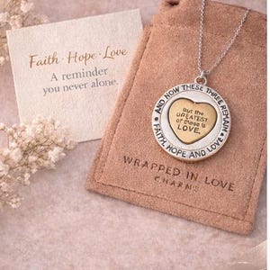 May include: A silver necklace with a heart-shaped pendant, inscribed with "But the GREATEST of these is LOVE." The pendant is on a chain, displayed with a card that reads "Faith Hope Love" and a reminder message. The necklace is in a tan pouch.
