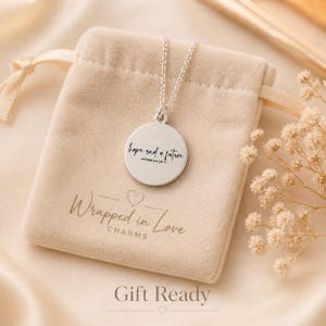 May include: A silver pendant necklace with a round charm engraved with the words "hope and a future" and the reference Jeremiah 29:11. The necklace rests on a beige velvet pouch with the text "Wrapped in Love Charms". The words "Gift Ready" are at the bottom.