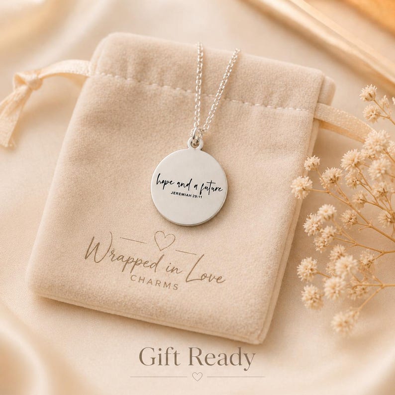 May include: A silver pendant necklace with a round charm engraved with the words "hope and a future" and the reference Jeremiah 29:11. The necklace rests on a beige velvet pouch with the text "Wrapped in Love Charms". The words "Gift Ready" are at the bottom.