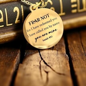 May include: A gold-toned circular pendant necklace featuring the engraved text FEAR NOT for I have redeemed you I have called you by name you are mine Isaiah 43:1. The pendant is suspended from a matching gold-toned metal chain link necklace.