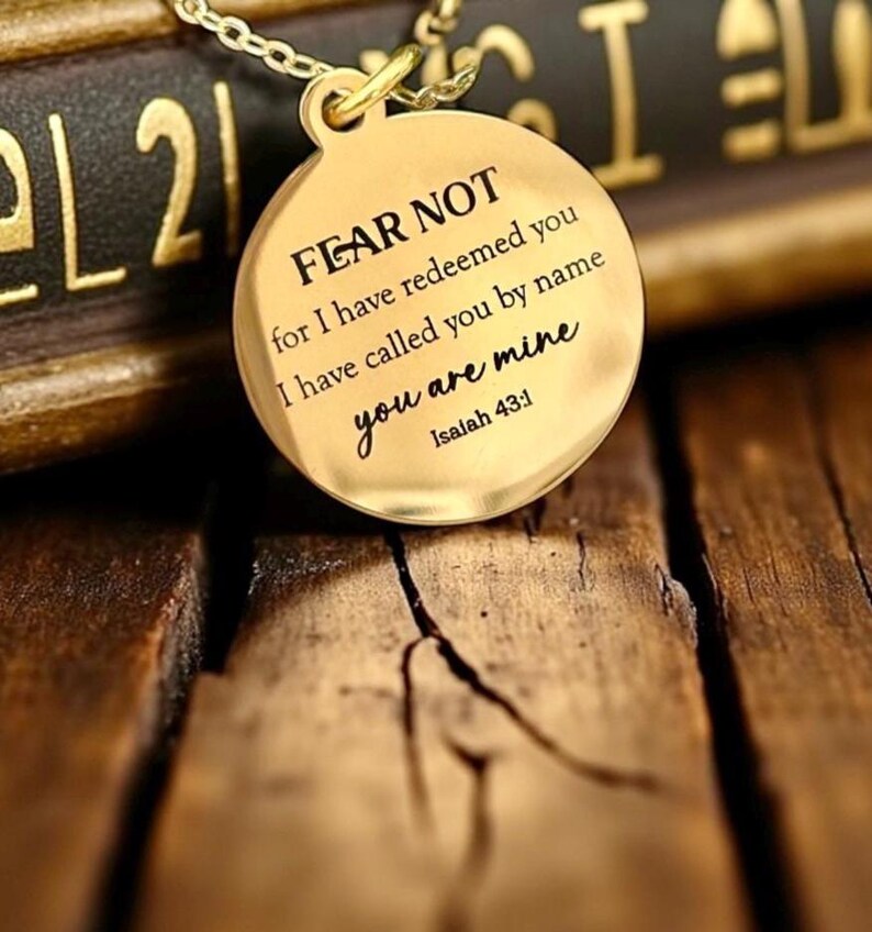 May include: A gold-toned circular pendant necklace featuring the engraved text FEAR NOT for I have redeemed you I have called you by name you are mine Isaiah 43:1. The pendant is suspended from a matching gold-toned metal chain link necklace.