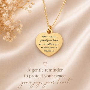 Op de afbeelding: Een goudkleurige hartvormige hanger aan een ketting met de gegraveerde tekst: Above all else guard your heart for everything you do flows from it PROVERBS 4:23. De hanger hangt aan een fijne goudkleurige ketting op een zachte stof.