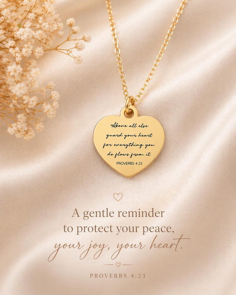 Op de afbeelding: Een goudkleurige hartvormige hanger aan een ketting met de gegraveerde tekst: Above all else guard your heart for everything you do flows from it PROVERBS 4:23. De hanger hangt aan een fijne goudkleurige ketting op een zachte stof.