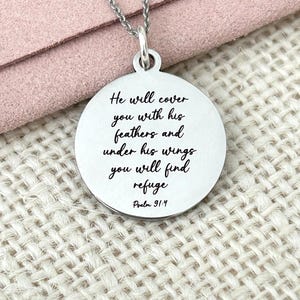May include: A silver pendant necklace featuring a circular charm. The charm is inscribed with the words "He will cover you with his feathers and under his wings you will find refuge Psalm 91:4" in black script.